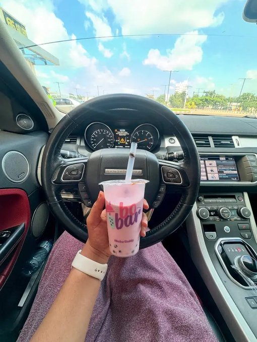 The image shows a person holding a pink drink with a straw, likely a smoothie or milkshake, while sitting inside a car. There are also other cars visible outside of the vehicle, suggesting that they might be at an event or gathering. The photo is related to the post because it features a pink drink and a car interior, both of which could be associated with the brand "Gm" in the image description.