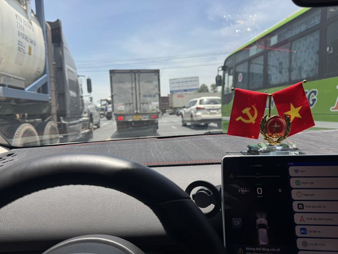 The image features a car with its dashboard screen visible, displaying some information related to traffic conditions outside of the vehicle. The car owner has chosen to display two Vietnamese flags as centerpieces on their dashboard rather than using traditional GPS directions or other typical road signs. This choice of decoration reflects an appreciation for the culture and language associated with Vietnam while possibly showing off a sense of patriotism as well. In addition, there are various notifications visible in the dashboard that may include information about traffic conditions, messages from friends, and even photos. The car's interior design elements serve to personalize it and create a unique atmosphere within the vehicle.