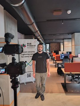 A man standing in a recording studio next to a camera on a tripod, with a banner behind him reading 'RGB Protocol - Bitcoin security, Lightning speed for every coin'.