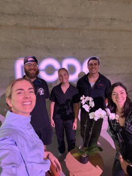 A group of five people pose for a selfie in front of a "NODE" sign, with a white orchid plant in the foreground. The caption celebrates the team's gathering at the NODE Foundation office in Palo Alto, highlighting the collaboration with @WAPSHOP_ETH, @karatekid_xyz, and @philmohun on bringing literary arts to the area, which the author jokingly calls "la tierra prometida."