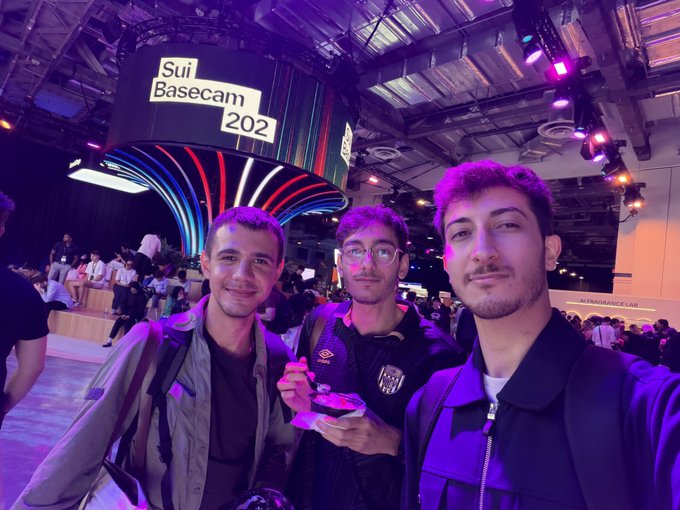 Three men take a selfie in front of a 'Sui Basecam 202' sign at what appears to be a conference or event space with purple lighting.