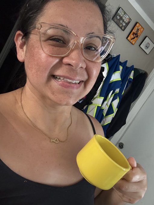 A smiling woman with glasses holds a yellow mug, wearing a necklace with a name pendant.