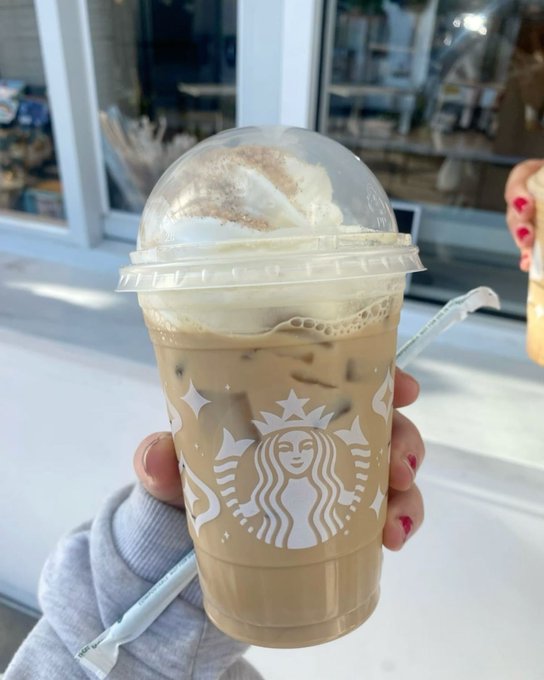 A hand with pink nail polish holding a clear plastic Starbucks cup containing an iced coffee drink topped with whipped cream and cinnamon, with a green paper straw.