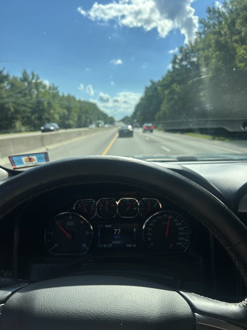 A driver's perspective photo from inside a car on the highway, with the speedometer showing 77 mph and the dashboard displaying "77" in the center, perfectly matching the post's mention of cruise control set to a target of $LINK. The image cleverly uses the 77 mph speed as a visual stand-in for the $LINK price target, with the open road and sunny sky ahead reinforcing the "on route to NC" journey.