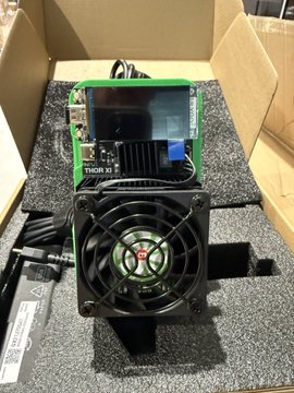 A photograph of cryptocurrency mining hardware in a cardboard box, featuring a green circuit board labeled 'TIDR X3' mounted with a large black cooling fan with red accents, sitting on gray foam packaging.