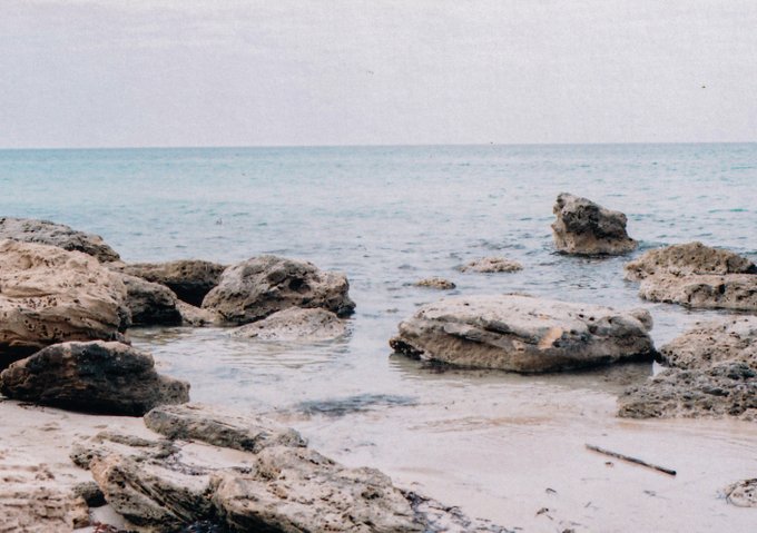 A serene coastal scene featuring large weathered rocks scattered across a sandy beach with calm turquoise water in the background, evoking a perfect "rather be here" Sunday getaway. The peaceful shoreline offers a dreamy escape from a day of outdoor chores.