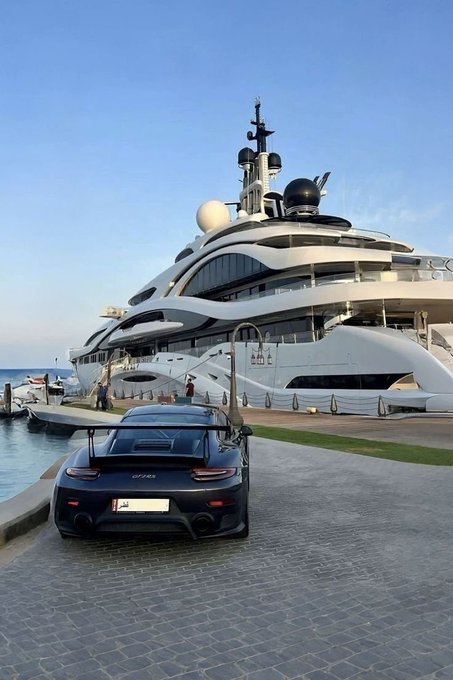 A black Porsche 911 GT2 RS is parked on a cobblestone dock beside a massive white superyacht, with the text "AM" visible on the yacht's side. The image juxtaposes two ultimate luxury status symbols to satirize the post's claim that achieving wealth is simply a matter of "larp[ing] your way to the top."