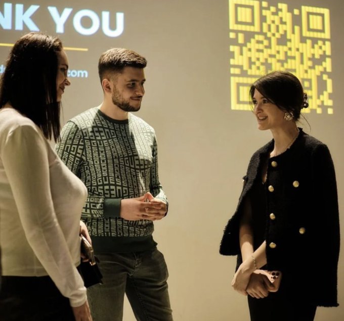 Three people conversing in front of a screen displaying a large yellow QR code and the partial text 'NK YOU' and '.com'.