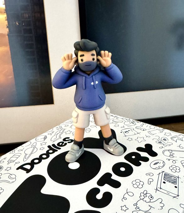 A 3D Doodles vinyl figure wearing a blue hoodie, gray shorts, and sneakers stands playfully on its branded "Doodles" and "VICTORY" packaging, matching the excitement of receiving a new collectible.