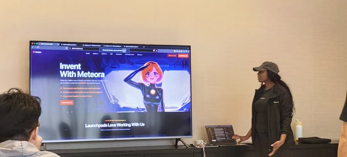 A woman in a black outfit and cap presents in front of a large screen displaying a webpage titled 'Invent With Meteora' featuring an anime-style character, with 'Launchpads Love Working With Us' at the bottom.