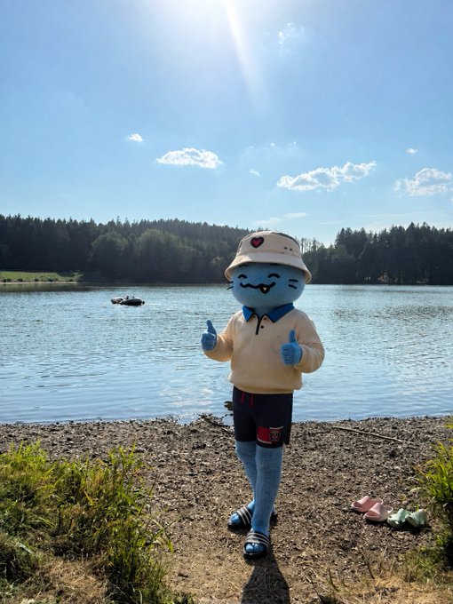 A blue mascot character wearing a cream-colored hoodie and a bucket hat, posing with a thumbs up on a sandy beach beside a lake.