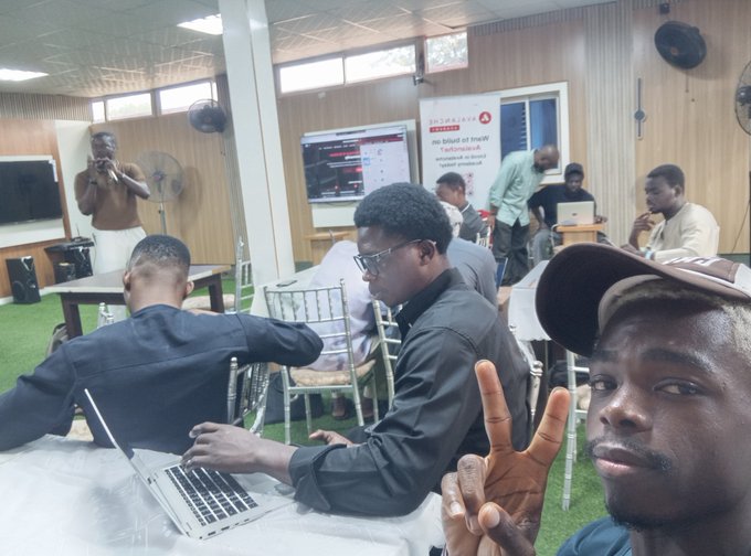 A group of young men work in a classroom or computer lab setting; one man in the foreground wears a cap and flashes a peace sign at the camera, while others work on laptops in the background near a screen displaying 'SatoshiLab', 'Telegram', and 'Decentralized (X)' for 'Bitcoin' and 'Ethereum'.
