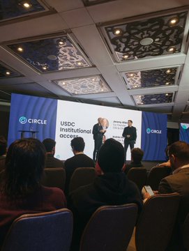 A conference room scene with two people presenting on stage to an audience, with screens displaying 'CIRCLE' and 'USDC Institutional accelerating'.