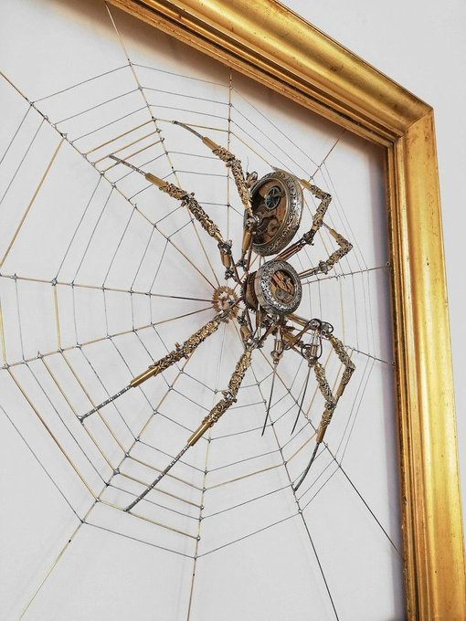 A framed steampunk-style spider sculpture crafted from antique watch gears, brass clockwork components, and intricate metallic legs, mounted against a white background with a delicate web of golden threads radiating outward. The piece is displayed within an ornate gilded frame, blending mechanical artistry with arachnid form.