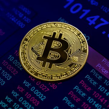 A physical Bitcoin coin rests on a dark background displaying financial data, with numbers like "10147" and "00000" visible, reflecting the cryptocurrency's price consolidation.