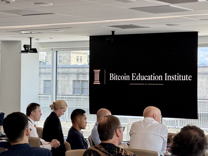 An audience seated in a modern conference room watching a presentation by the 'Bitcoin Education Institute' displayed on a large screen.