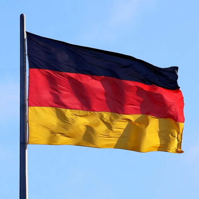 The image shows the German flag (black, red, and gold horizontal stripes) waving on a flagpole against a blue sky, symbolizing Germany in relation to the news about the country's planned 25% cryptocurrency capital gains tax starting in 2027.