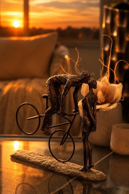 A bronze sculpture of two figures sharing a tender kiss—one on a bicycle, leaning down to meet the other—sits on a stone base, bathed in the warm golden glow of a sunrise through the window.

The image beautifully complements the post's message of pausing from the markets to enjoy a peaceful mountain morning, as the soft sunrise lighting and intimate sculpture evoke a serene, reflective start to the day away from the charts.