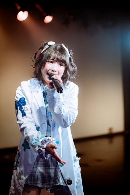 A young woman performing on stage, singing into a microphone while wearing a white jacket with blue star designs, a plaid skirt, and white hair ribbons.