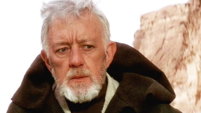 A close-up of an older man with white hair and a beard wearing a brown hooded robe, resembling Obi-Wan Kenobi from Star Wars.