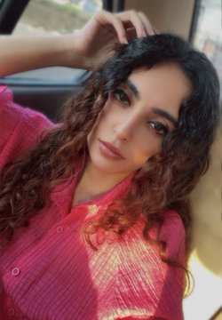 A young woman with long, curly dark hair poses for a selfie, wearing a bright pink textured shirt while seated inside a car.