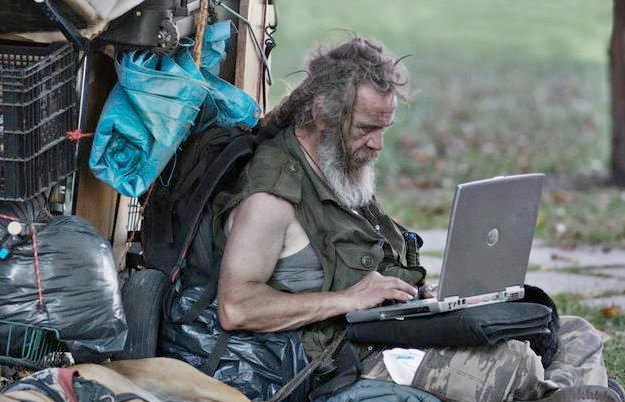 A disheveled older man with a long beard sits outdoors using a silver laptop, surrounded by belongings including blue plastic bags and black trash bags.