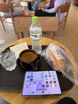 A tray on a cafe table holds a foldable smartphone displaying app icons, a cup of coffee, a bottle of water, and a wrapped pastry.