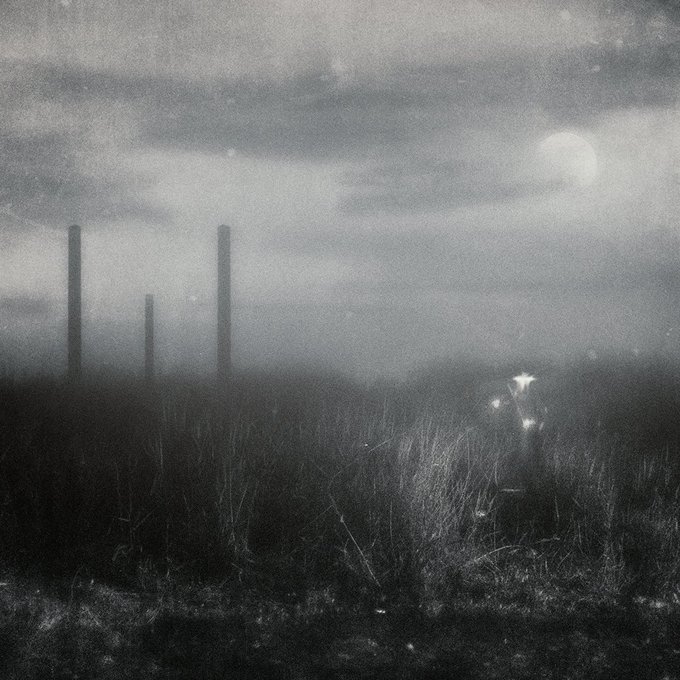 A misty, eerie black-and-white landscape featuring industrial smokestacks in the distance, a luminous moon, and a faint figure with glowing lights in a grassy field.