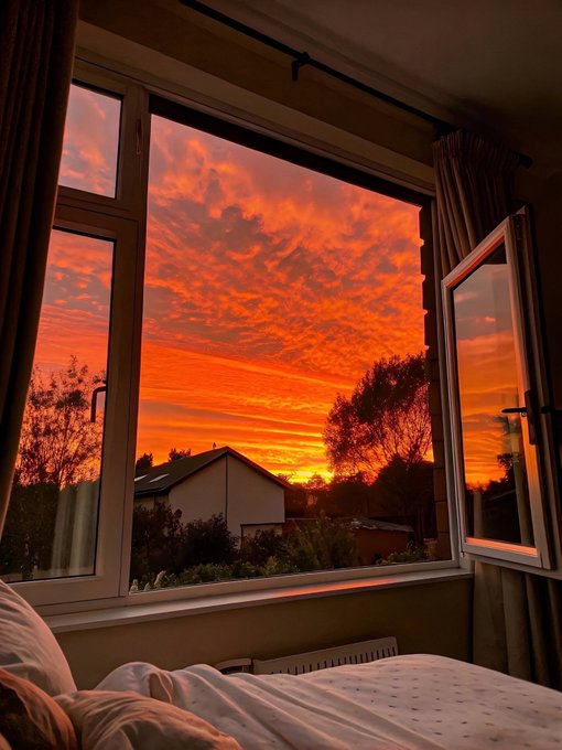 A bedroom view through an open window showing a dramatic, vibrant orange and red sunset over trees and rooftops.
