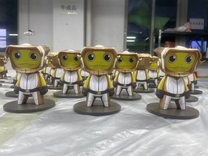 A row of assembled papercraft frog figures in white and gold astronaut-style suits displayed on a workshop table with Chinese text '半成品' (semi-finished product) visible in the background.