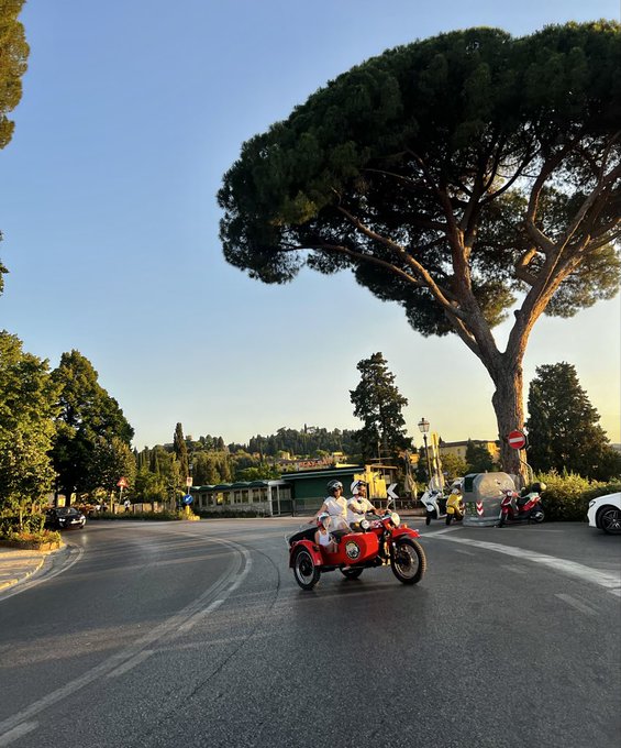 Two people ride a red motorcycle with a sidecar around a curved road beneath a large pine tree in a scenic European-looking setting.