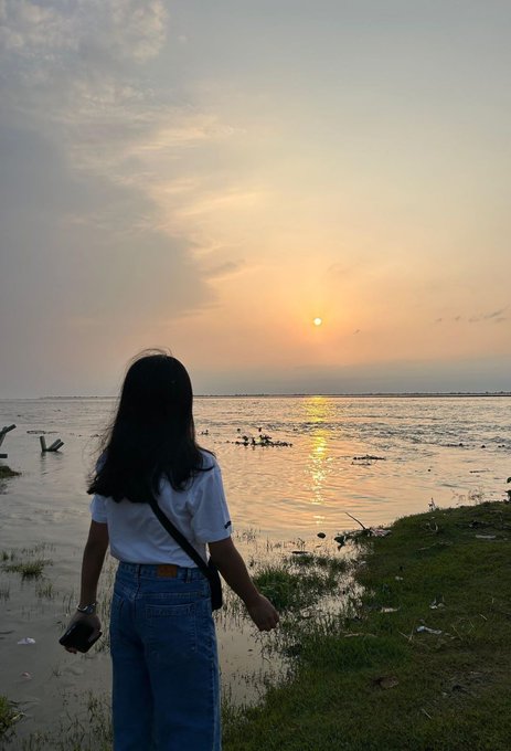 A scenic photograph of a young woman with long dark hair standing by a body of water at sunset, viewed from behind, wearing a white shirt and blue jeans while holding a phone.