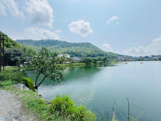 Photograph of a scenic lake surrounded by lush green hills, trees, and a few buildings under a partly cloudy blue sky.