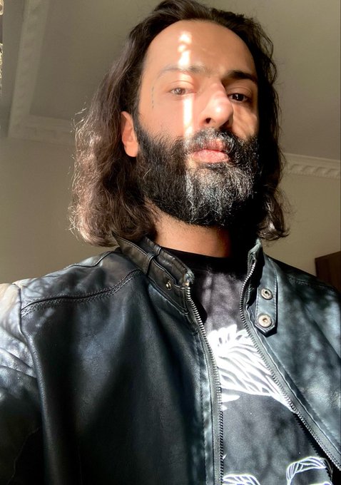A selfie-style photo of a bearded man with long dark hair wearing a black leather jacket over a graphic t-shirt, with a strip of sunlight visible on his face.