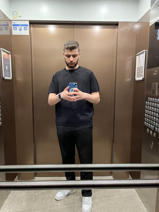 A man taking a mirror selfie in an elevator wears a black t-shirt, black pants, and white sneakers while looking at his smartphone.