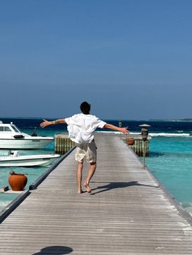 A man walks barefoot down a wooden pier extending over turquoise water, with his arms outstretched to his sides and boats visible to the left.