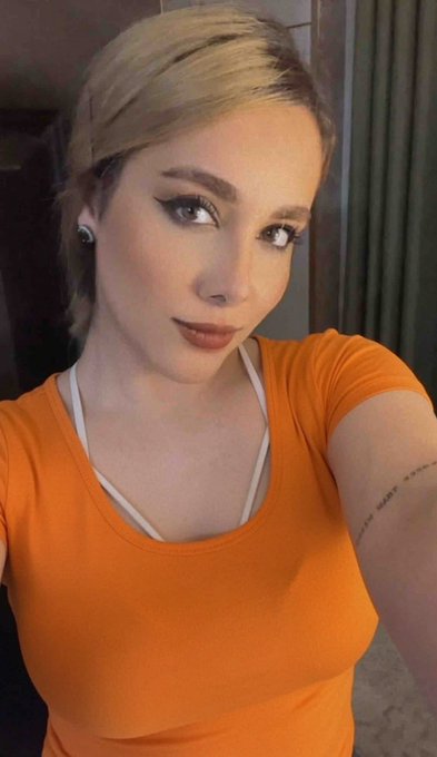 A blonde woman with winged eyeliner takes a selfie while wearing a fitted orange t-shirt with a white strap visible underneath.