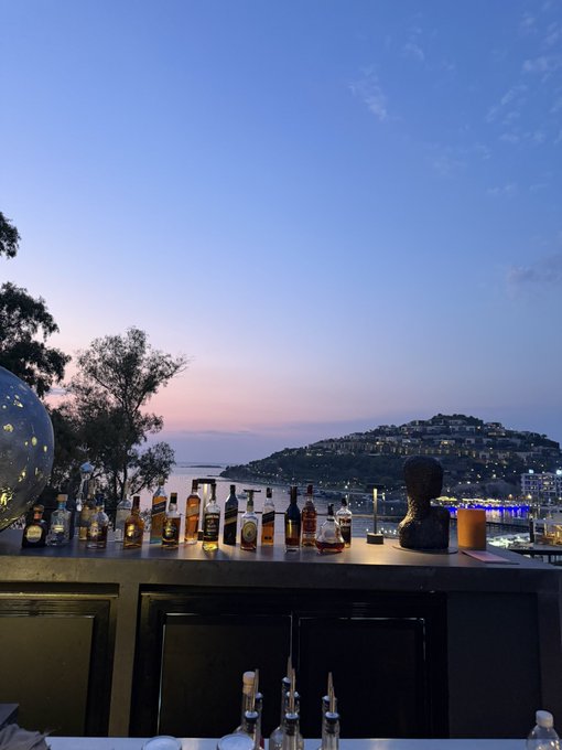 A row of liquor bottles lines an outdoor bar overlooking a coastal hillside town at dusk under a pink and blue sunset sky, with "JACK DANIEL'S" visible on one of the labels. The serene evening view pairs perfectly with the casual "Gm" (good morning) greeting, offering a peaceful start-of-day ambiance.