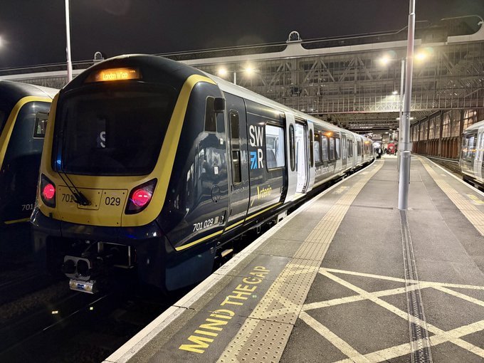 A dark blue and yellow SWR train with the number 702029 idles at a nighttime station platform marked with "MIND THE GAP".