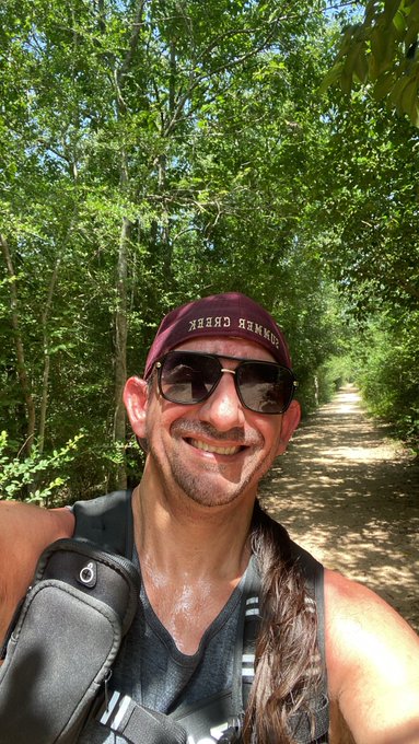 A smiling man wearing sunglasses and a "GREEN BERET" cap takes a selfie on a sun-dappled forest trail while hiking with a hydration pack, embodying the post's message of taking action—his sweat and forward movement through the woods visually reinforcing the "Get After It" mantra of building momentum through doing.
