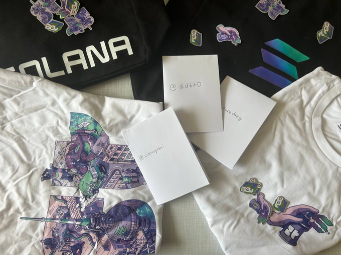 The image features a collection of merchandise, including two shirts with unique designs - one white shirt adorned with purple and green illustrations and another black shirt featuring a cartoon character. The shirts are accompanied by a note that reads "solana" on the white shirt and "superteam" on the black shirt. Additionally, there is a postcard in the image, which includes text such as "sending merch and handwritten letters to three different continents that'@solana and @superteam culture". The items are arranged on a table or countertop, with each item placed next to its corresponding description.
