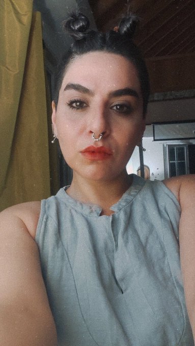 Selfie of a woman with dark hair styled in space buns, wearing a septum piercing and a light blue sleeveless top in a home setting.