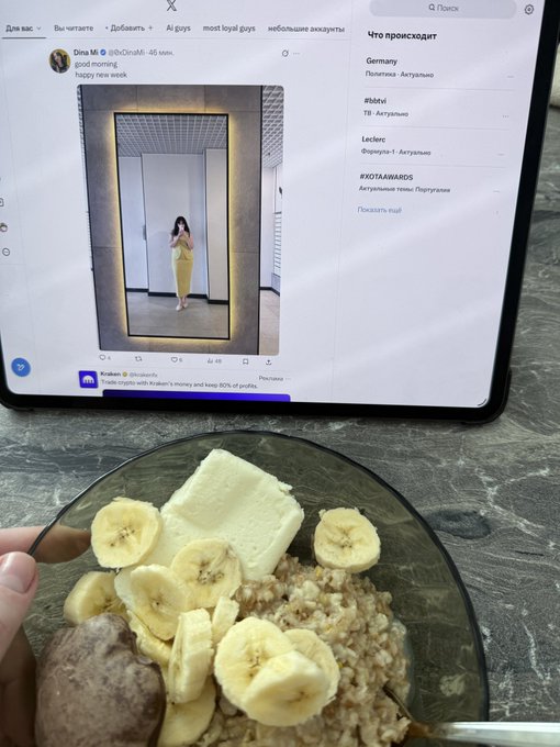 A bowl of oatmeal topped with banana slices and butter sits in front of a tablet displaying an X post with a mirror selfie and Russian text, including “tbvi,” “Germany,” and “#otv4.”