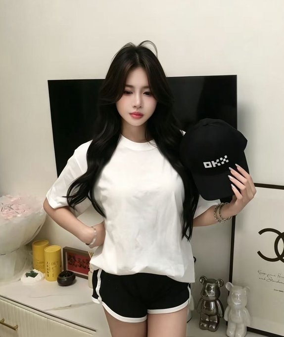 A young woman with long dark hair wearing a white t-shirt and black shorts with white trim stands indoors holding a black cap with white 'OKX' lettering, with Chanel-themed decor visible in the background.