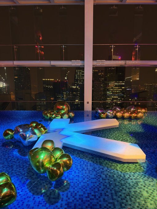 A nighttime rooftop party with blue-lit mosaic tiles features an inflated white Twitter "X" logo surrounded by clusters of metallic gold and iridescent balloons, with a city skyline visible in the distance—likely the venue for the exclusive KAST.xyz dinner where Elon Musk's rebranding of Twitter to "X" is being celebrated or questioned.