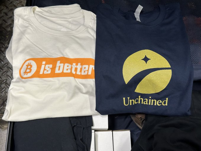 Two folded t-shirts displayed side by side: a white one reading '₿ is better' in orange and a navy one with 'Unchained' and a yellow logo.