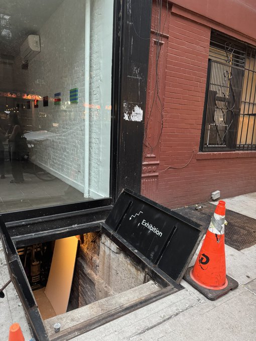 An open sidewalk hatch marked "Exhibition" reveals a subterranean gallery entrance beside a red brick building, framed by an orange traffic cone—visually reinforcing the post's argument that meaningful culture happens below street level, not within institutional white cubes.