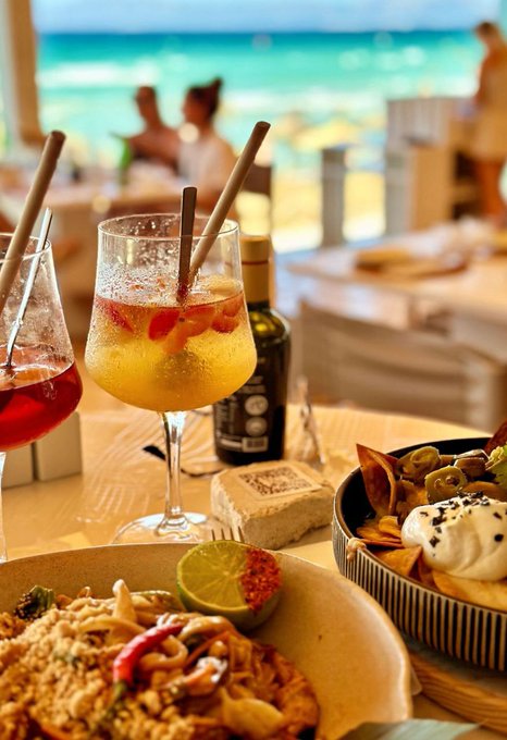 A coastal brunch spread features an Aperol spritz and a Limoncello bottle alongside dishes of pasta with lime, a flatbread with burrata, and olives, all set against a turquoise sea view that perfectly matches the celebratory weekend vibe of making $5,000+ in July.
