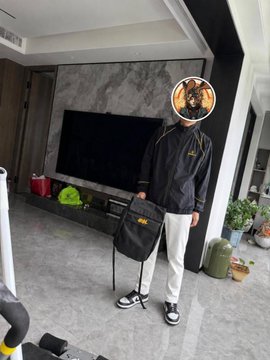 A person wearing a black Binance jacket, white pants, and black sneakers stands holding a black messenger bag, with their face covered by a Binance logo graphic. The caption mentions a "Binance 9th Anniversary Gift Box" airdrop, and the person is showcasing the gift box attire in their post.
