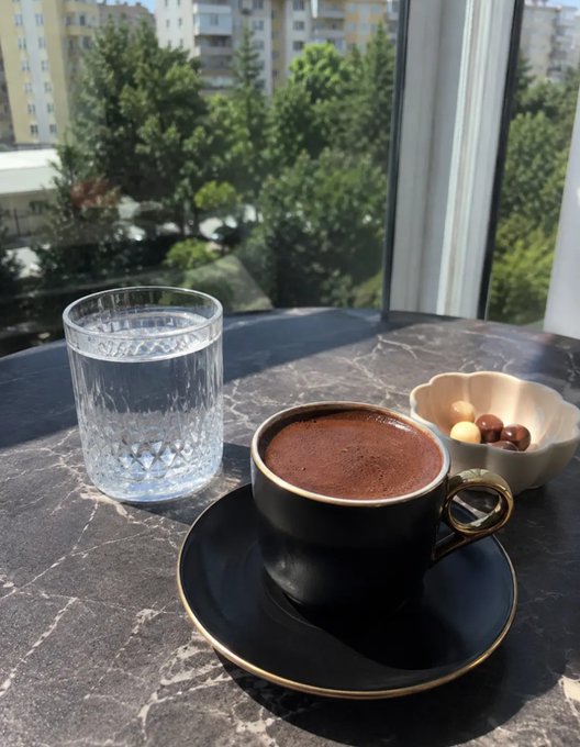 A black coffee cup with gold trim on a saucer, a glass of water, and a small bowl of colorful round candies sit on a dark marble table near a window overlooking green trees and city buildings.